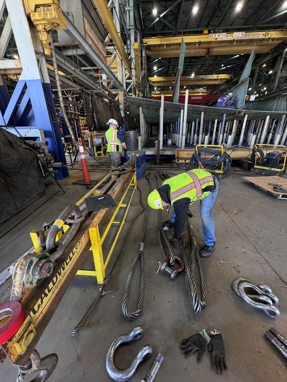 Nelson inspectors checking wire rope slings and a spreader beam in a shipyard, under overhead cranes