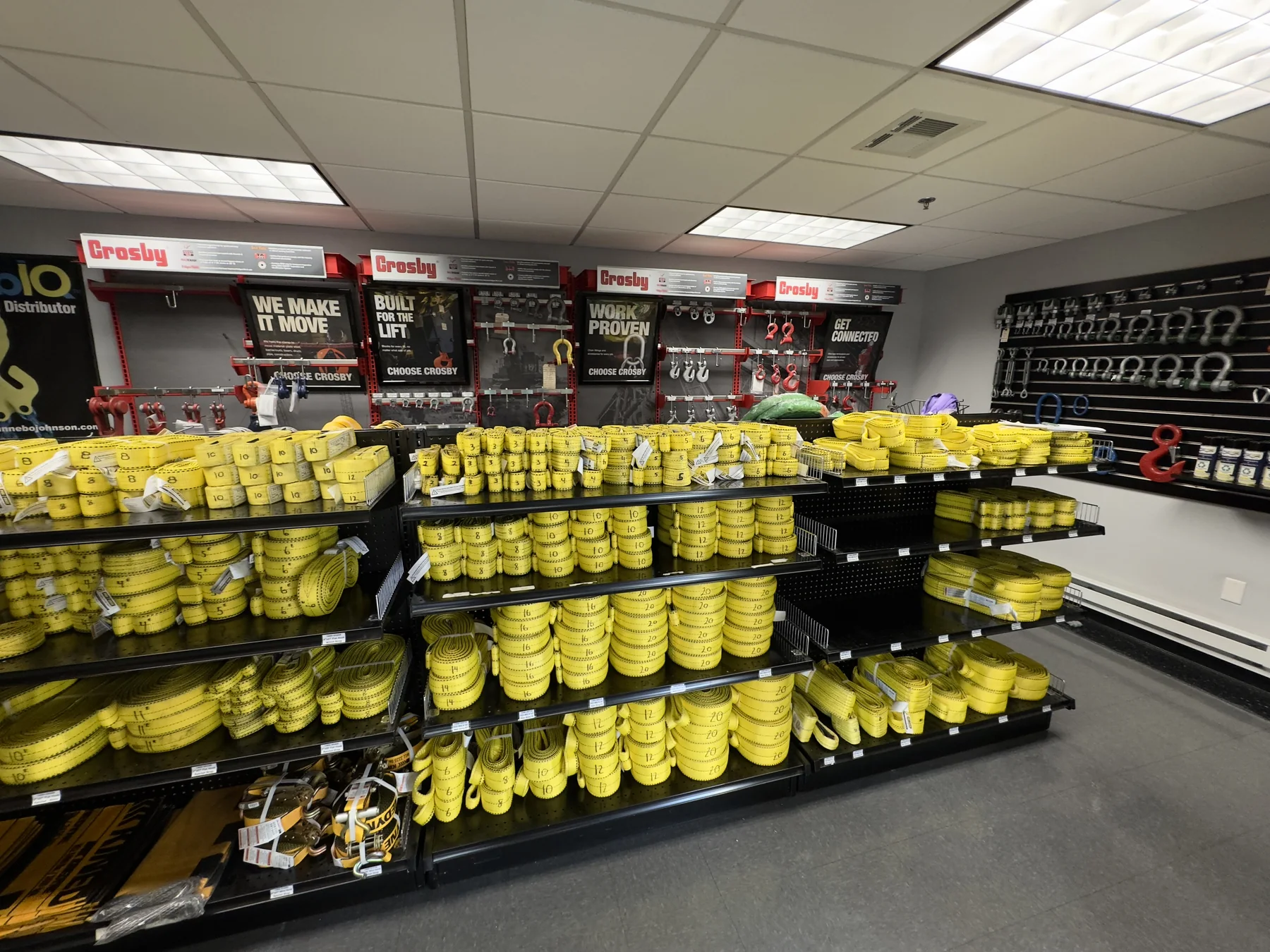 Showroom shelves of yellow web slings sorted by width and length, with Crosby hardware displays and shackles and hooks hung on pegboard behind