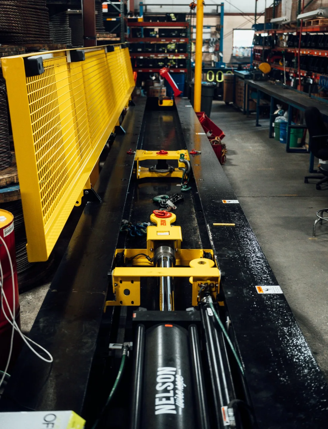 Looking down the length of the test bed: the hydraulic ram and its yellow carriage in the foreground, a master link and chain rigged along the bed, shop racking either side