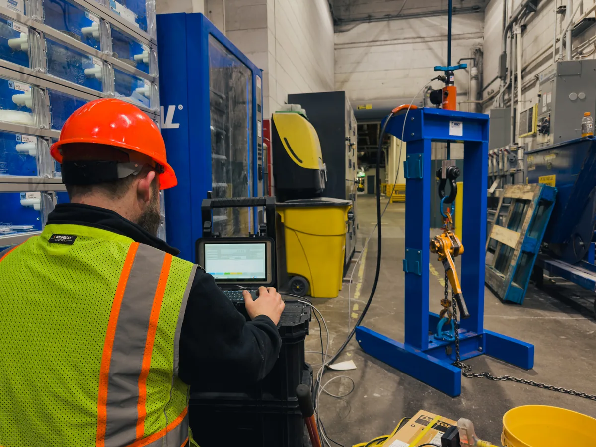 A Nelson technician running a proof test on site: a portable blue test frame with a lever hoist and chain rigged in it, load logged live on a laptop