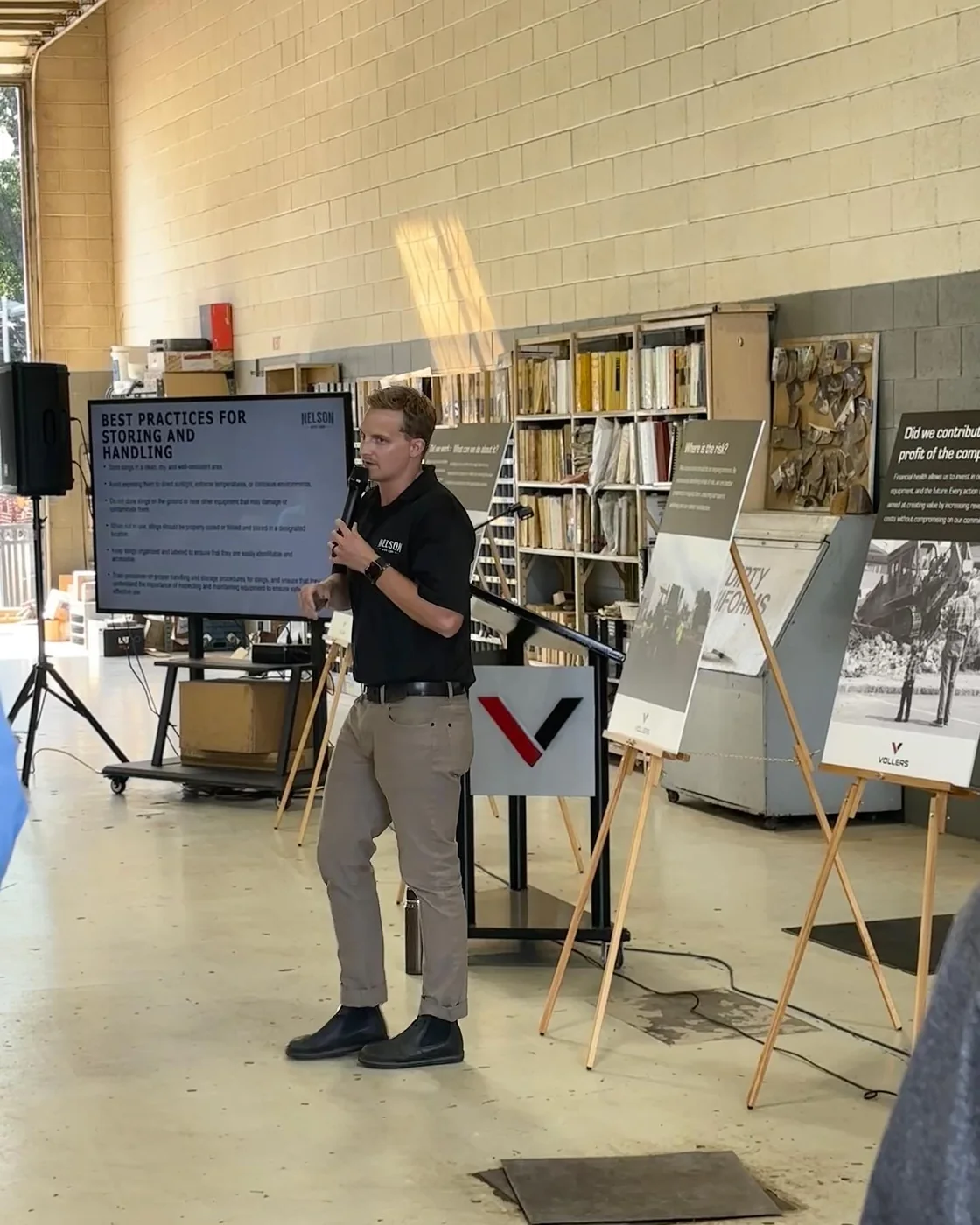 Steve Nelson speaking with a microphone at a customer safety day, beside a screen showing a Nelson Wire Rope slide titled Best Practices for Storing and Handling